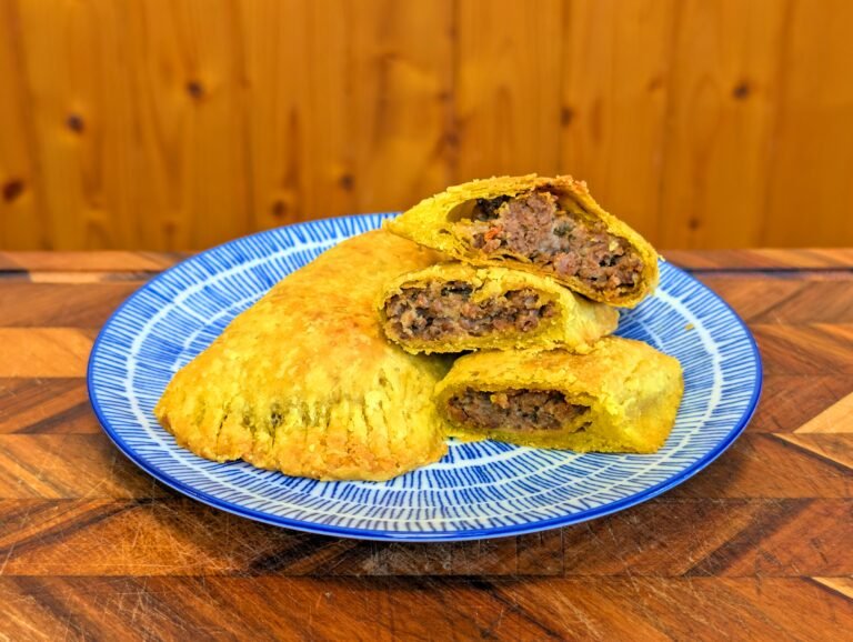 A blue and white patterned plate featuring one whole Jamaican beef patty and several halved pieces, revealing the moist, dark savory meat filling inside a flaky crust.