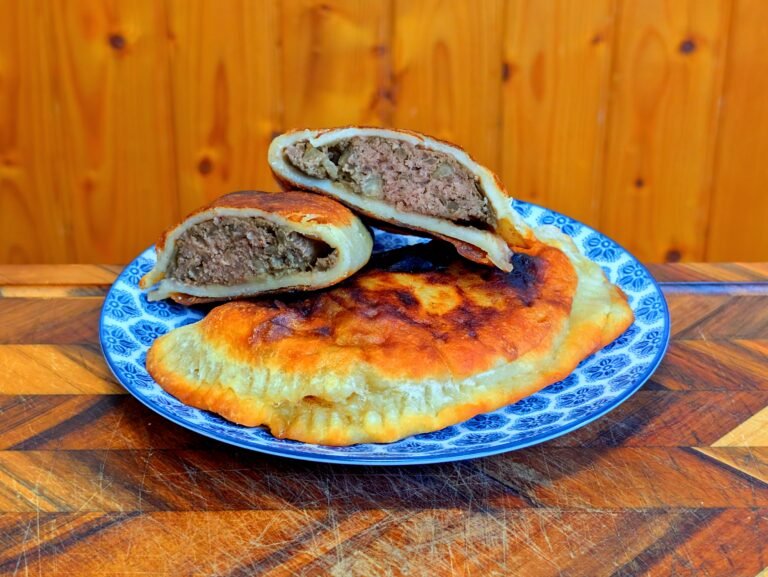 Close-up of freshly pan-fried chebureki on a blue and white patterned plate, with one cut in half to reveal a juicy ground meat and onion filling.