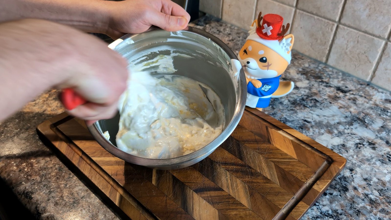 Hand using a red spatula to fold and mix a thick white frosting in a stainless steel bowl.