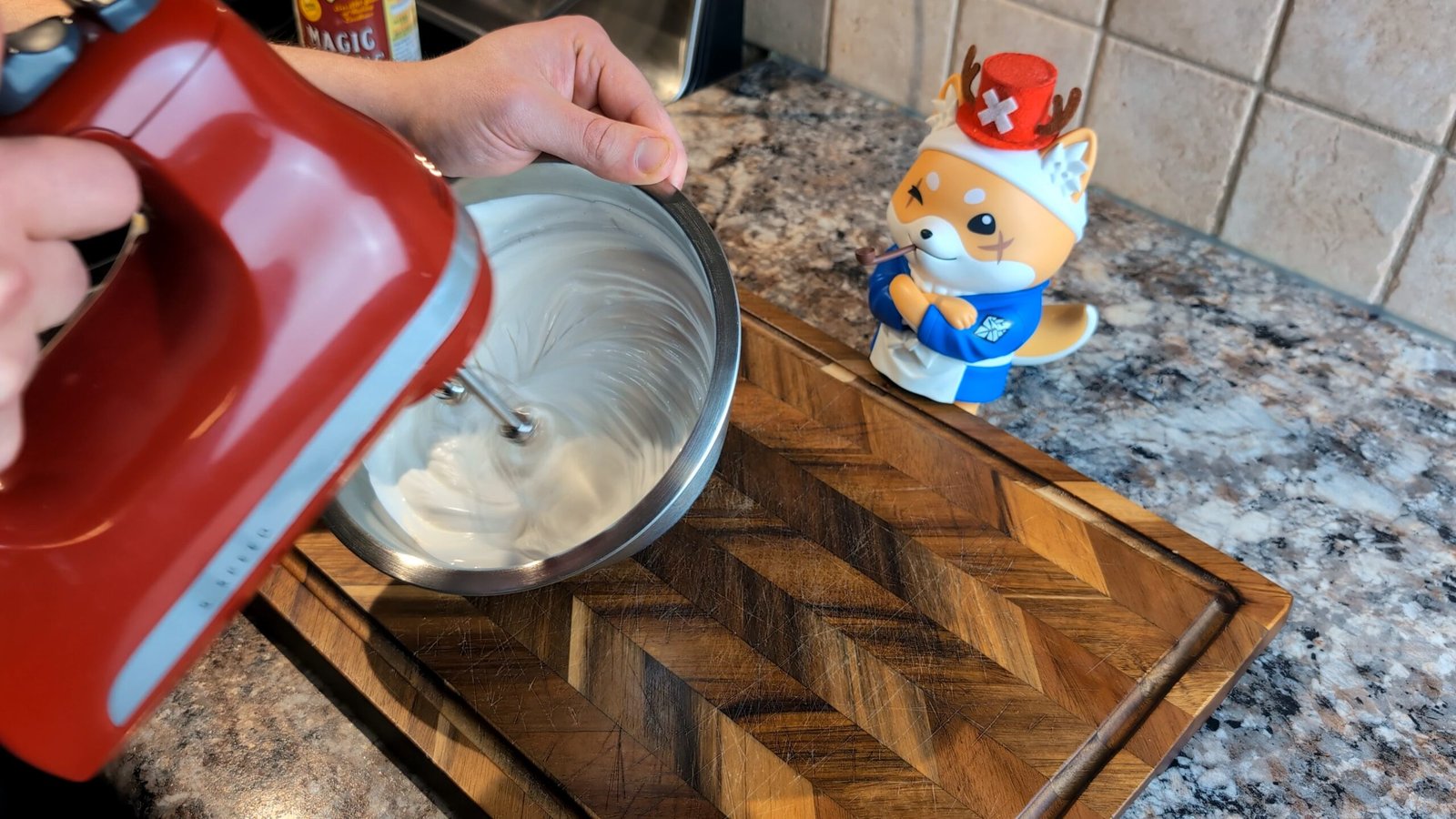 Hand using a red electric mixer to whip glossy white frosting in a stainless steel bowl next to a Shiba Inu figurine.
