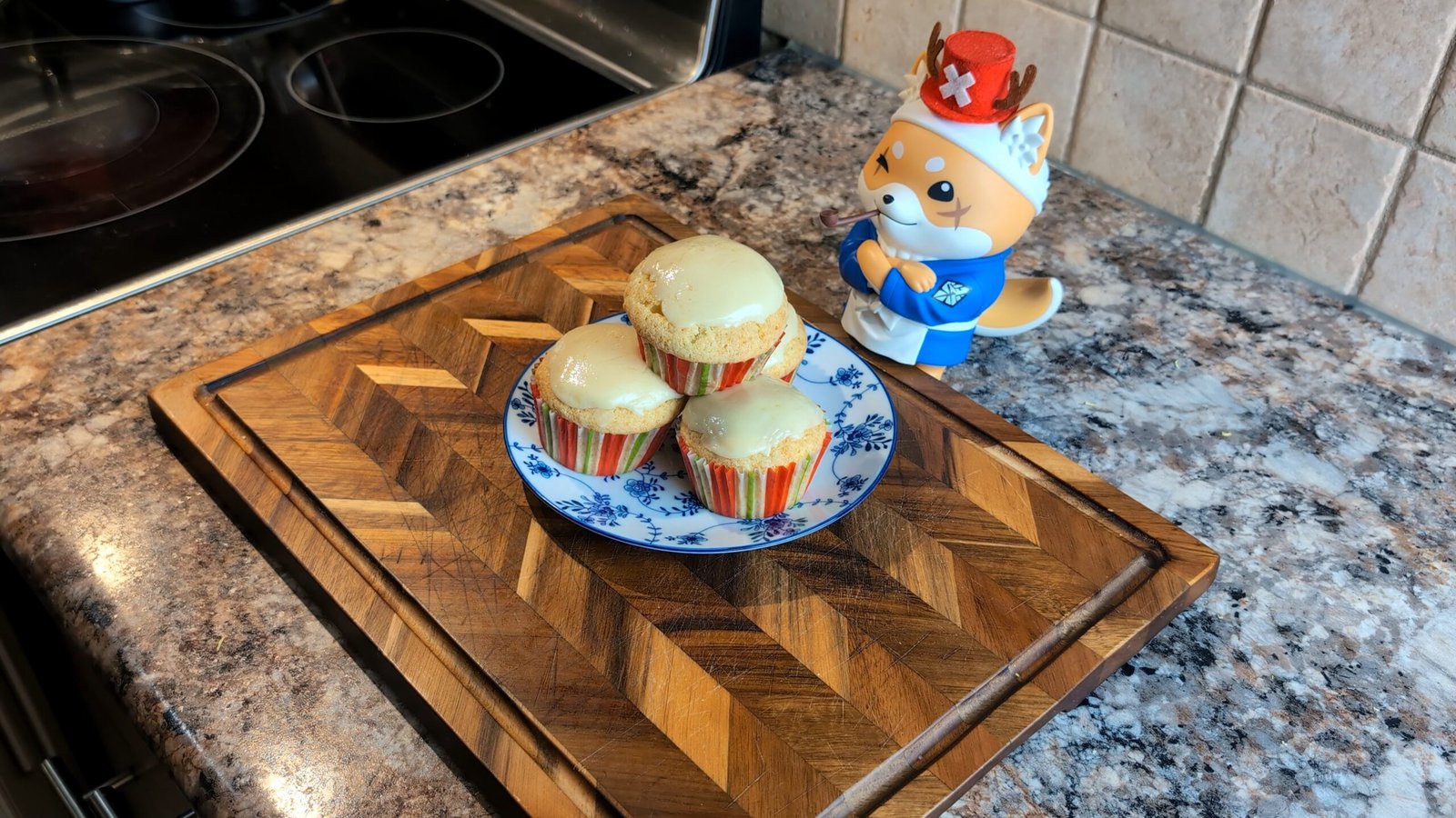 Three freshly glazed lemon cupcakes sitting on a wooden herringbone cutting board next to a Shiba Inu figurine.