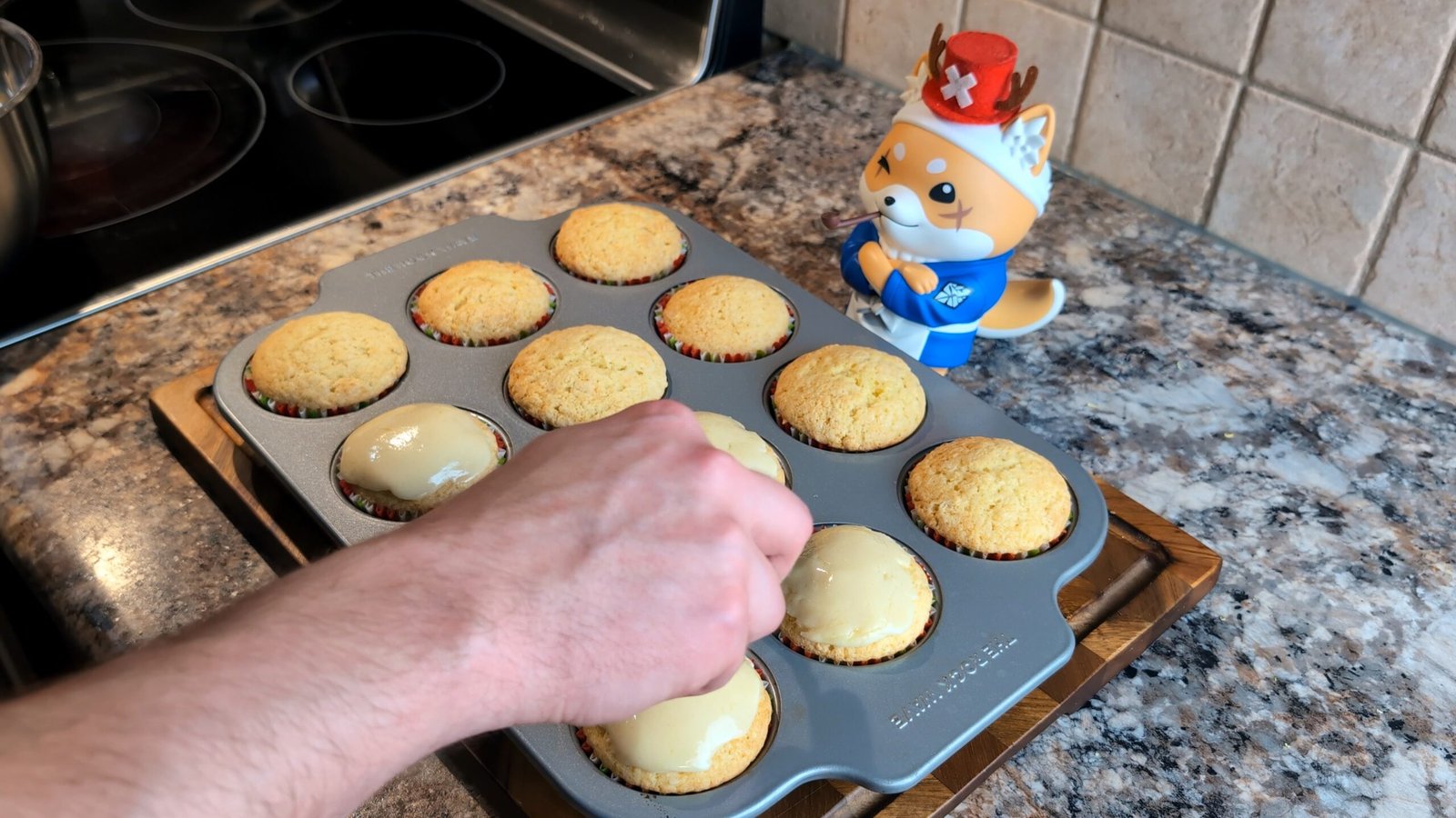 Hand dipping the top of a baked lemon cupcake into a smooth white glaze over a metal muffin tin.