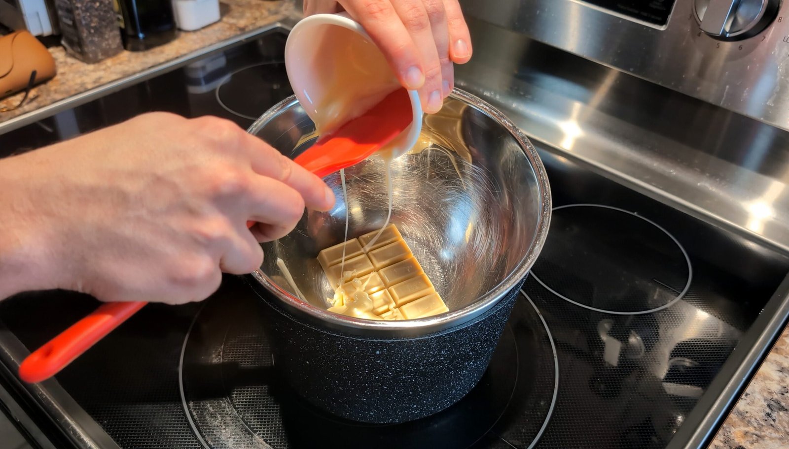 Pouring condensed milk over white chocolate squares in a metal mixing bowl set over a pot of simmering water to create a glaze.