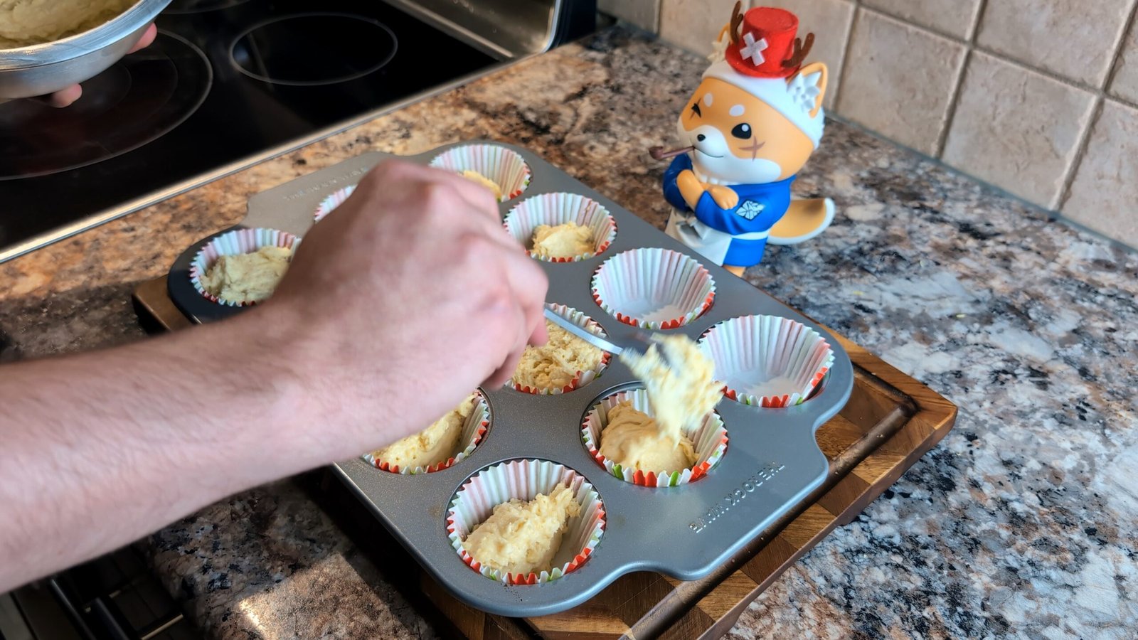 Hand using a spoon to scoop thick lemon cupcake batter into colorful paper liners inside a baking tin.