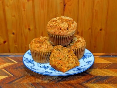 A stack of four freshly baked carrot muffins on a blue and white patterned plate, with one muffin broken open to reveal the moist interior dotted with grated carrots.