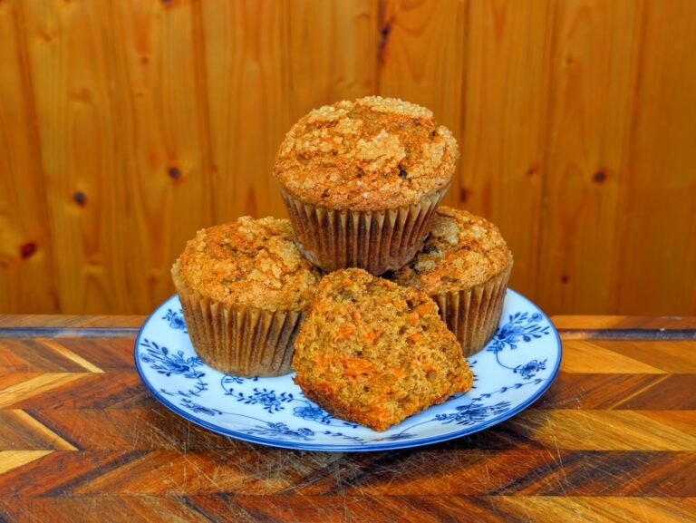 A stack of four freshly baked carrot muffins on a blue and white patterned plate, with one muffin broken open to reveal the moist interior dotted with grated carrots.