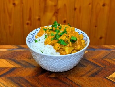 A decorative white bowl filled with creamy orange cauliflower butter masala and fluffy white rice, garnished with fresh chopped cilantro.