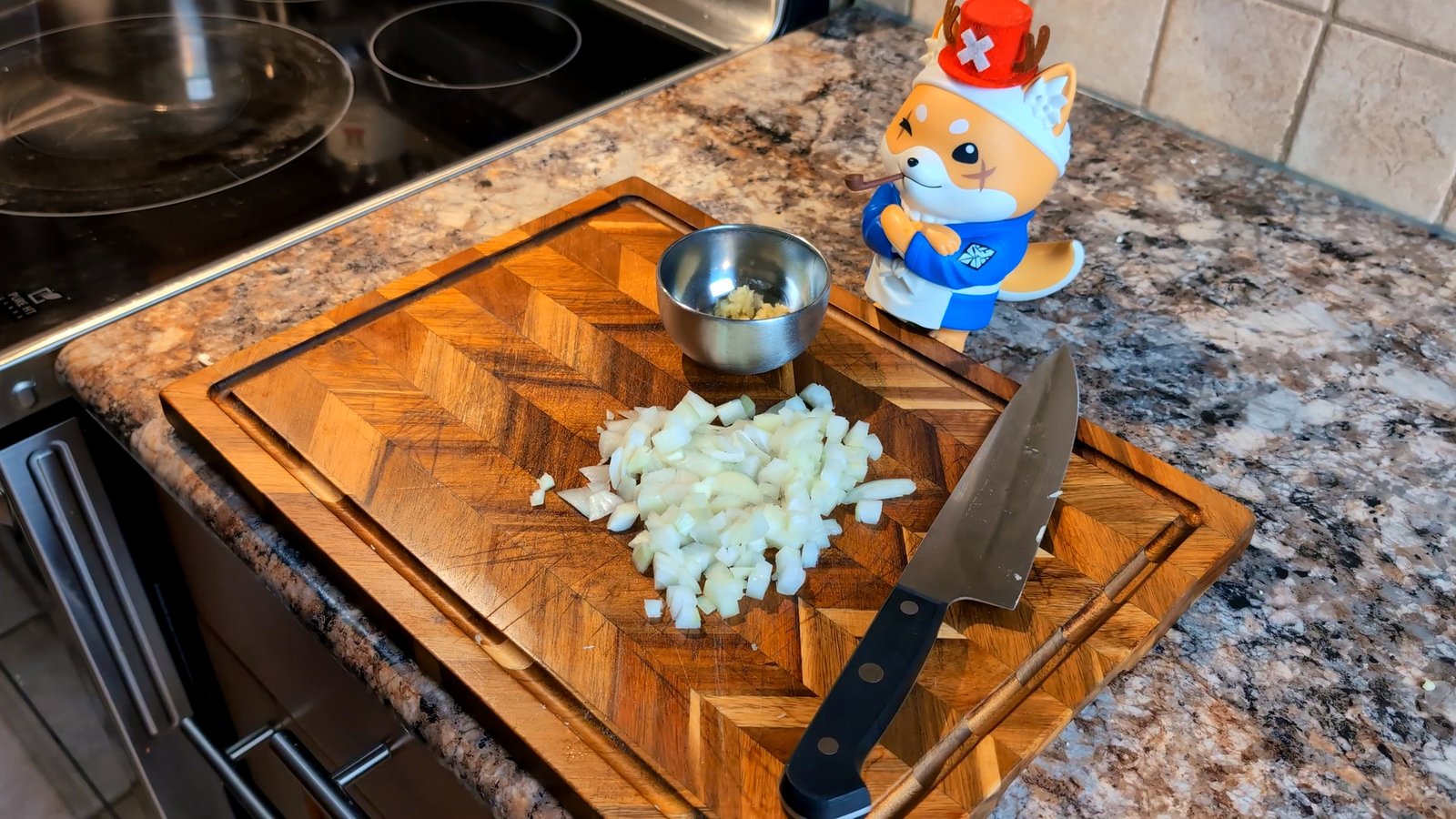 Finely diced white onions and a chef's knife on a wooden cutting board, next to a small metal bowl of minced garlic.