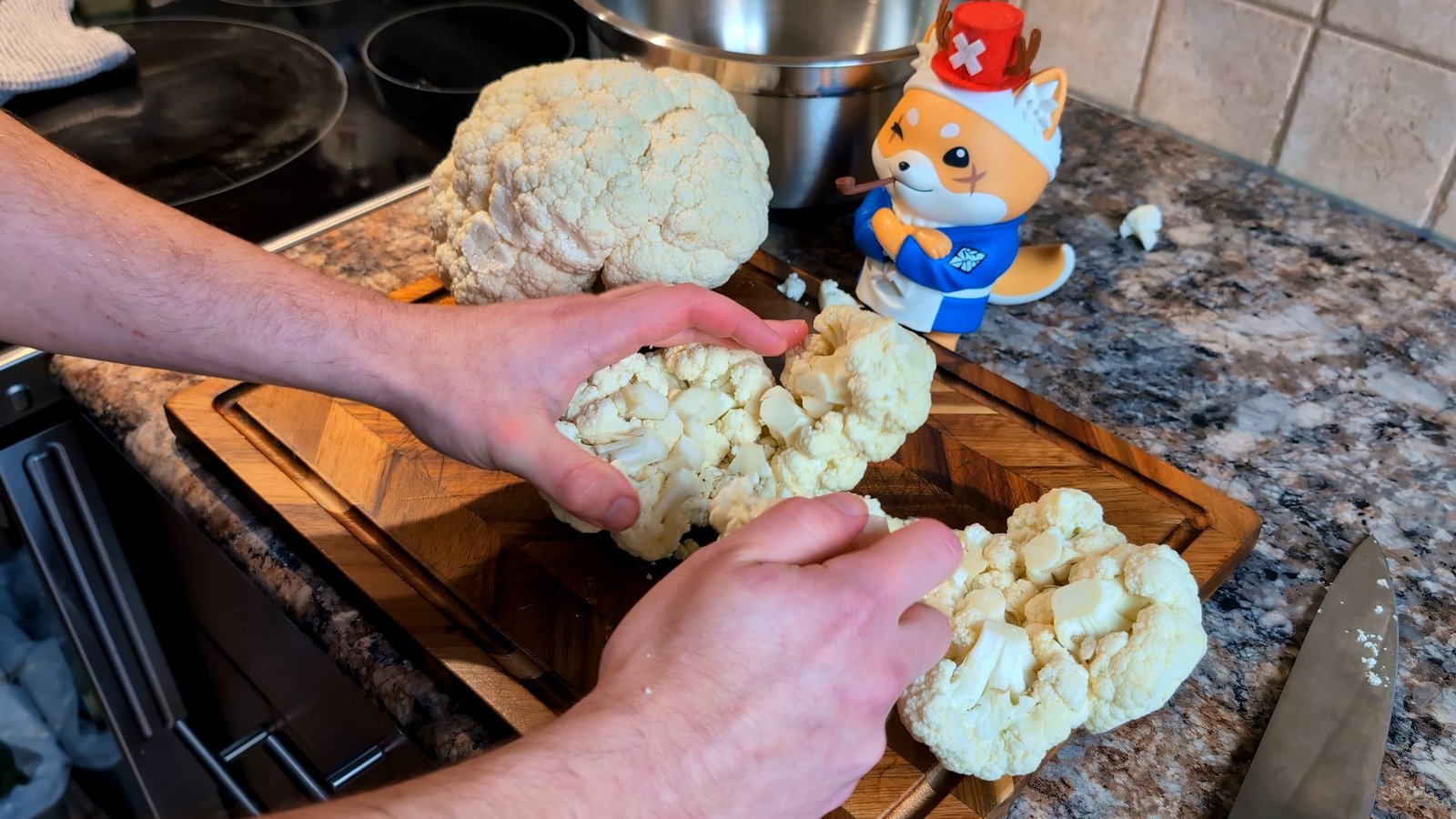 Two hands breaking apart a large, fresh head of raw cauliflower into smaller florets on a wooden cutting board.
