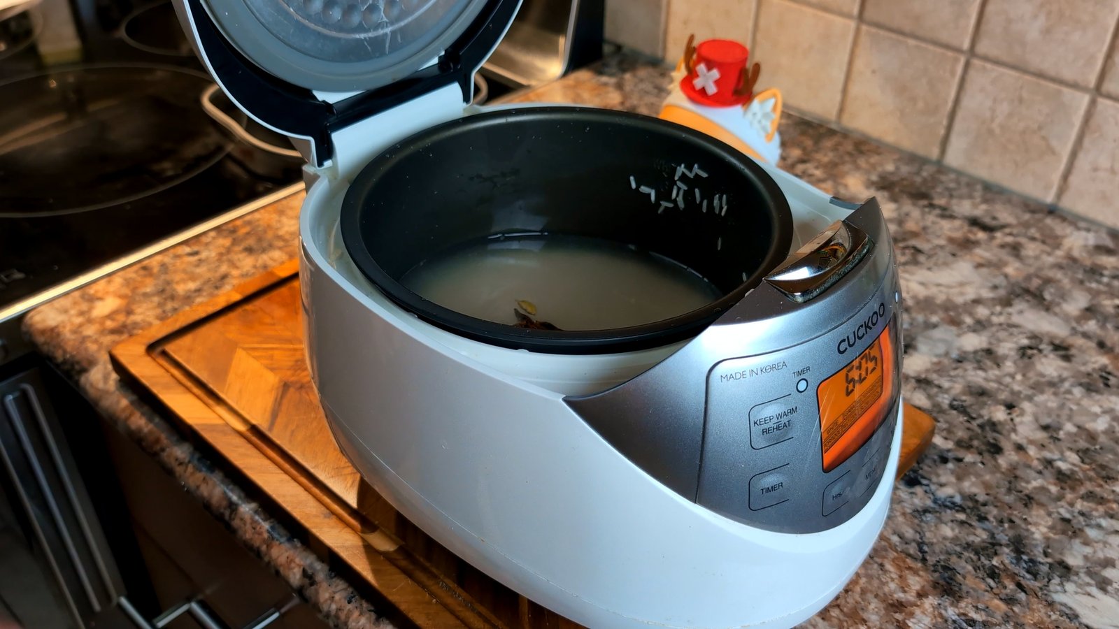 An open white Cuckoo brand rice cooker filled with water, uncooked white rice, and a few whole spices like cardamom pods.