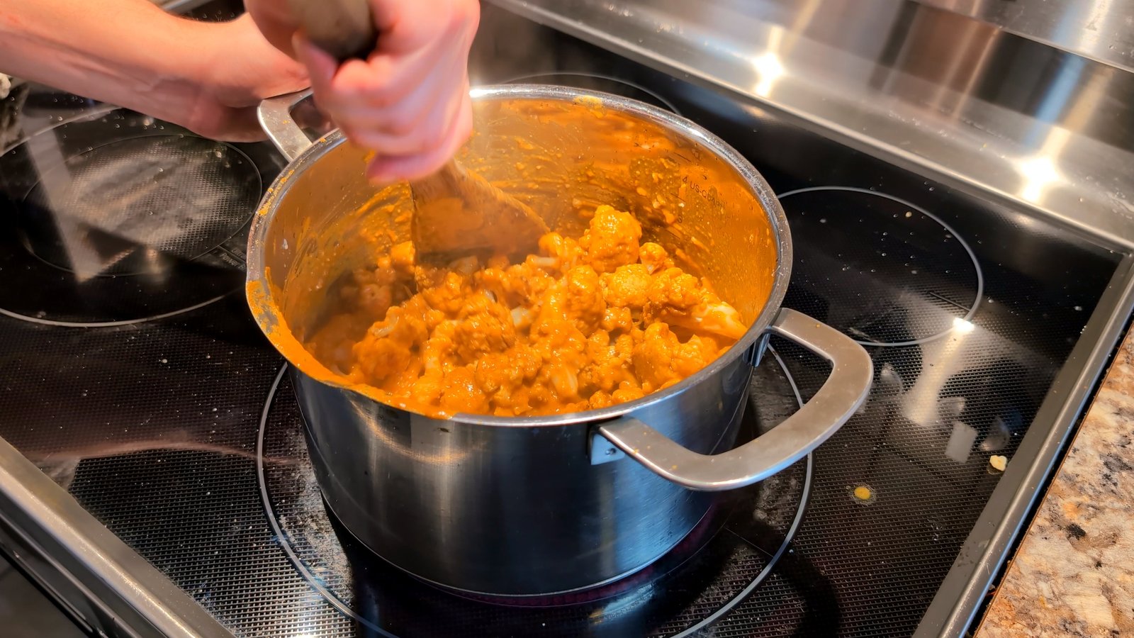 Stirring roasted cauliflower florets into a simmering, creamy orange butter masala sauce in a stainless steel pot.