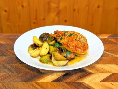 A white bowl filled with a golden-brown pan-seared chicken breast topped with minced garlic and fresh parsley, served alongside roasted halved Brussels sprouts and multi-colored fingerling potato wedges in a light herb sauce.