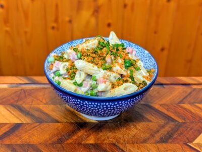 A blue and white patterned bowl filled with creamy penne pasta, peas, and ham, topped with golden toasted breadcrumbs and fresh parsley, set against a wooden background.