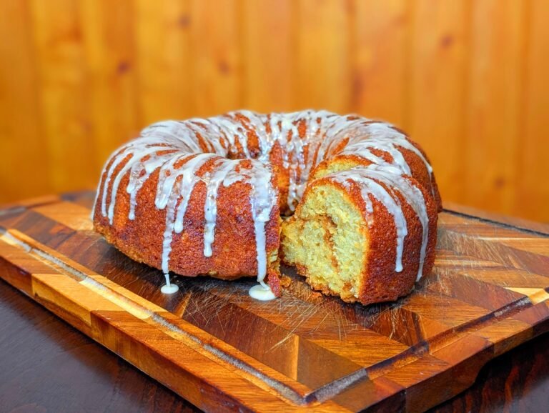 A close-up of a perfectly baked maple coffee cake on a wooden cutting board, generously drizzled with white glaze. A slice is pulled forward, revealing a thick, cinnamon-sugar streusel ribbon baked inside.