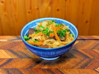 A finished bowl of Mongolian Tsuivan noodles served in a blue and white patterned ceramic bowl. The dish features thick, hand-cut noodles mixed with tender chunks of beef, julienned carrots, and onions, topped with a generous garnish of fresh chopped green onions.