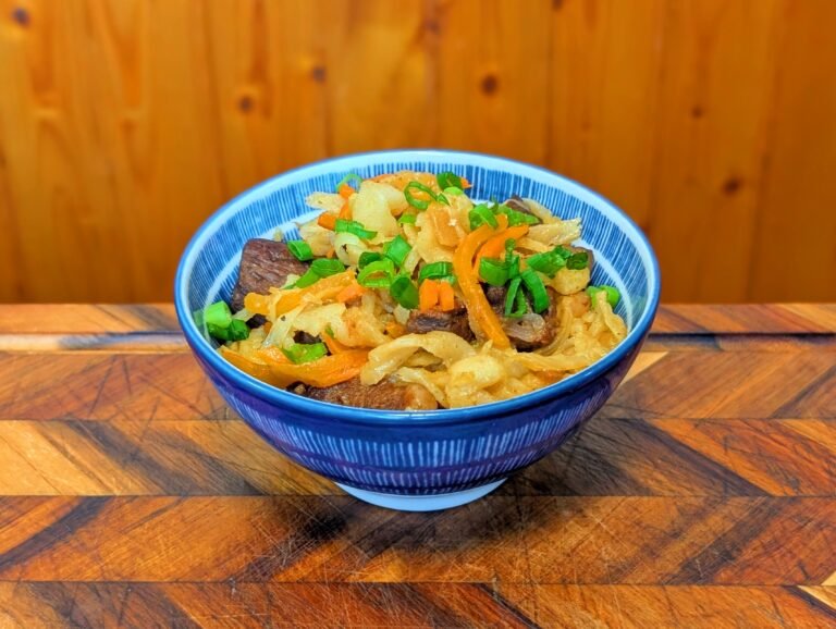 A finished bowl of Mongolian Tsuivan noodles served in a blue and white patterned ceramic bowl. The dish features thick, hand-cut noodles mixed with tender chunks of beef, julienned carrots, and onions, topped with a generous garnish of fresh chopped green onions.