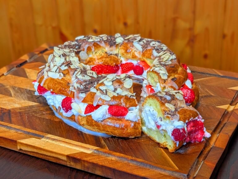A completed Strawberry and Raspberry Paris-Brest on a chevron wooden board with a slice removed, showcasing the thick interior layer of whipped cream, fresh strawberries, and whole raspberries.